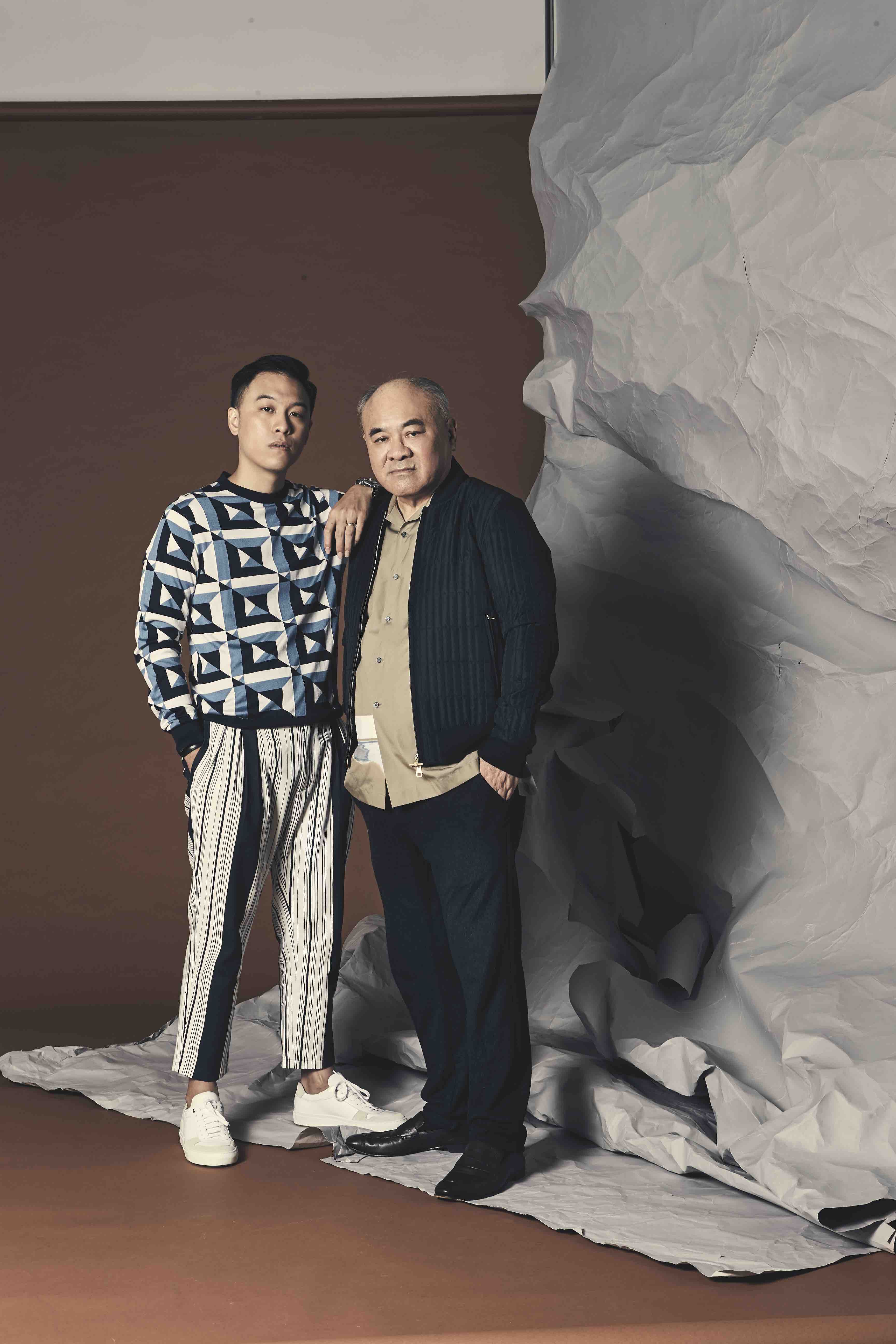 Jon is wearing a geometric print cotton knit sweater and  cotton striped trousers from Dolce & Gabbana, and white leather sneakers, from Boss. Jon's father is wearing a cotton short sleeved shirt and black quilted bomber jacket from Dunhill.