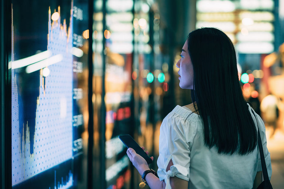 Young businesswoman checking financial trading data on smartphone by the stock exchange market display screen board in downtown financial district